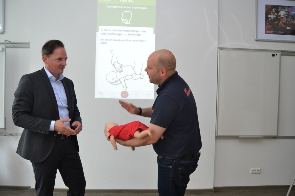 Christopher Tomaszik (re.) zeigt Barmer-Regionalgeschäftsführer Patrick Schäfer, was zu tun ist, wenn der Säugling einen Fremdkörper in der Luftföhre hat. Im Hintergrund gibt die App die Handlungsanweisungen. (Foto: Johanniter/ Sabine Eisenhauer)